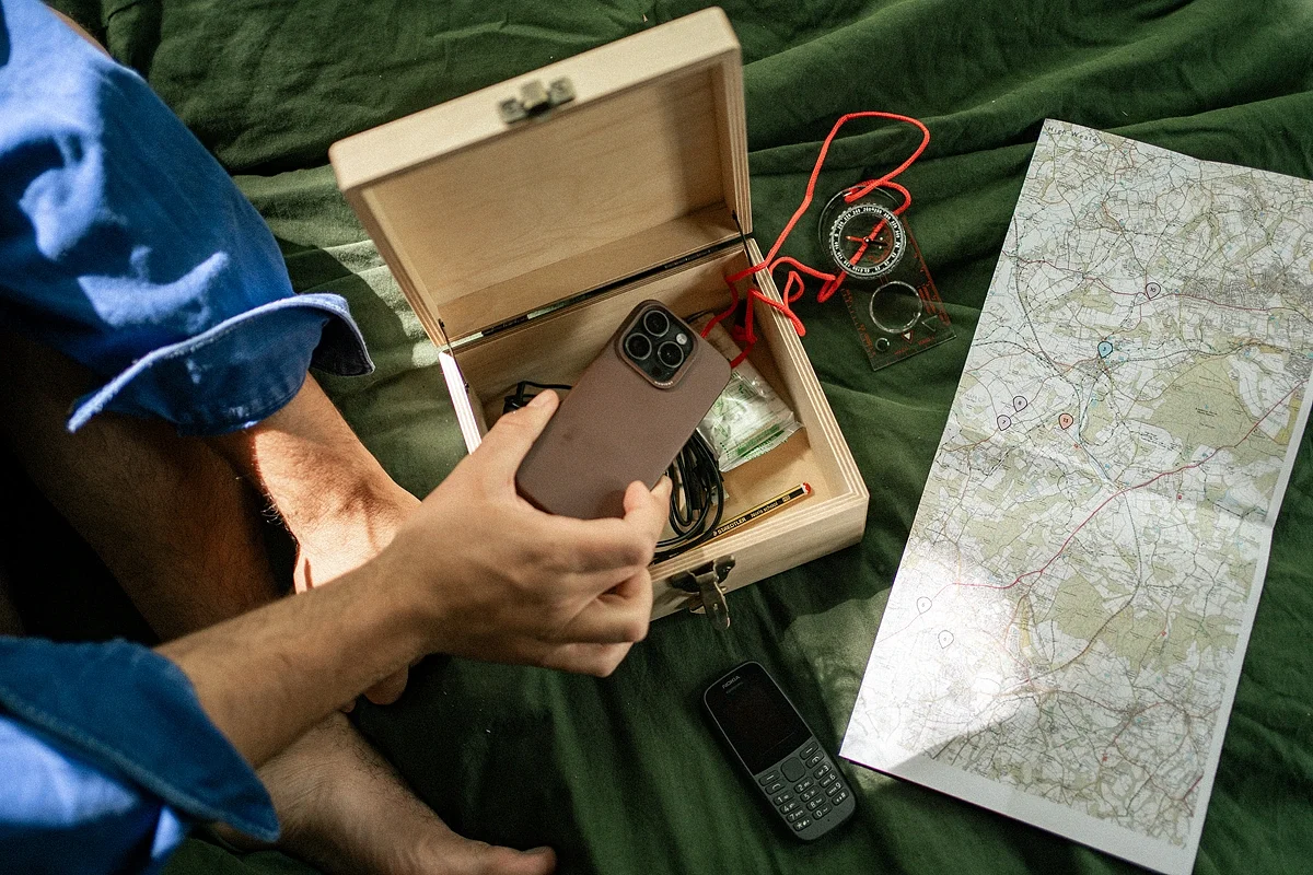 A hand placing a smartphone into an open wooden box, next to a basic phone, a compass and a paper map